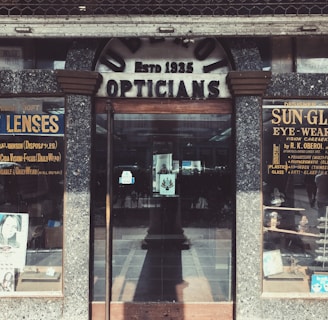 A vintage storefront for an optician, featuring weathered signage reading 'Opticians' and 'Estb 1935'. Display cases on either side showcase products like contact lenses and sunglasses, with advertisements for various eyewear brands. The building has a rustic, aged look with textured stonework surrounding the entrance.