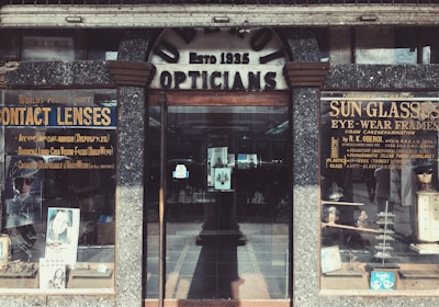 A vintage storefront for an optician, featuring weathered signage reading 'Opticians' and 'Estb 1935'. Display cases on either side showcase products like contact lenses and sunglasses, with advertisements for various eyewear brands. The building has a rustic, aged look with textured stonework surrounding the entrance.