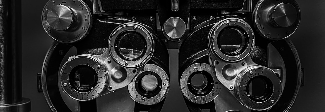 A close-up view of an optometry device called a phoropter, used for testing individual lenses on each eye during an eye exam. The device is composed of multiple dials, lenses, and knobs.