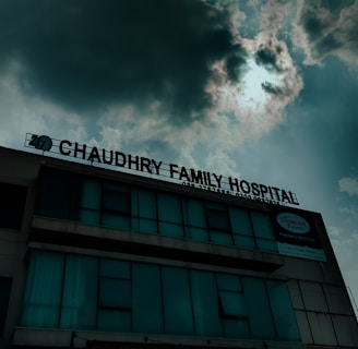 A hospital building with a sign that reads 'Chaudhry Family Hospital', positioned under a cloudy sky with visible sunlight peeking through. The building has multiple windows, some of which are covered or tinted.