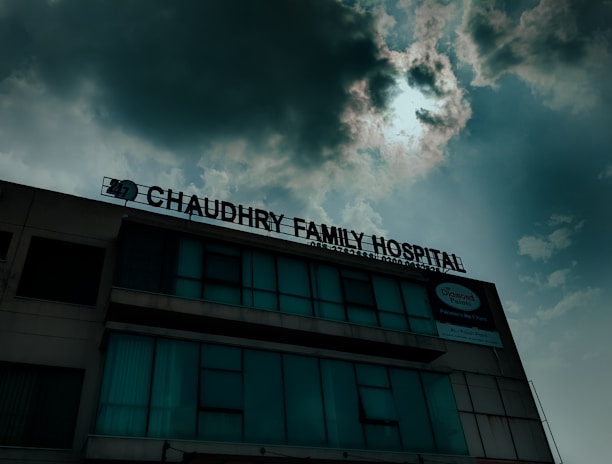 A hospital building with a sign that reads 'Chaudhry Family Hospital', positioned under a cloudy sky with visible sunlight peeking through. The building has multiple windows, some of which are covered or tinted.