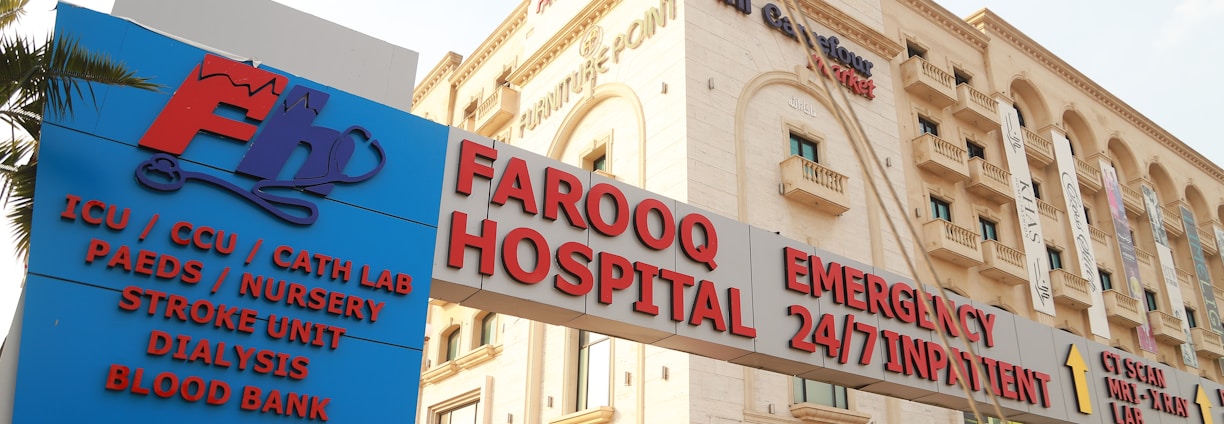 A hospital building with an entrance sign indicating various departments such as ICU, CCU, Cath Lab, Paediatrics, Nursery, Stroke Unit, Dialysis, and Blood Bank. The building has a beige facade with multiple stories and several windows. There are various signs with red and blue lettering highlighting emergency services and 24/7 inpatient care.