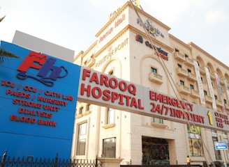 A hospital building with an entrance sign indicating various departments such as ICU, CCU, Cath Lab, Paediatrics, Nursery, Stroke Unit, Dialysis, and Blood Bank. The building has a beige facade with multiple stories and several windows. There are various signs with red and blue lettering highlighting emergency services and 24/7 inpatient care.