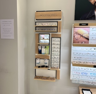 A display wall featuring multiple shelves with various eyeglasses and related accessories. The shelves are labeled 'ACCESSORIES' and include categories like 'COMPUTER GLASSES', 'READY READERS', and 'CLIP-ON FRAMES'. There are promotional materials and images of glasses on the wall, as well as a signboard thanking customers for shopping. A sign indicates the store is under CCTV surveillance.