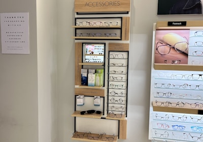 A display wall featuring multiple shelves with various eyeglasses and related accessories. The shelves are labeled 'ACCESSORIES' and include categories like 'COMPUTER GLASSES', 'READY READERS', and 'CLIP-ON FRAMES'. There are promotional materials and images of glasses on the wall, as well as a signboard thanking customers for shopping. A sign indicates the store is under CCTV surveillance.